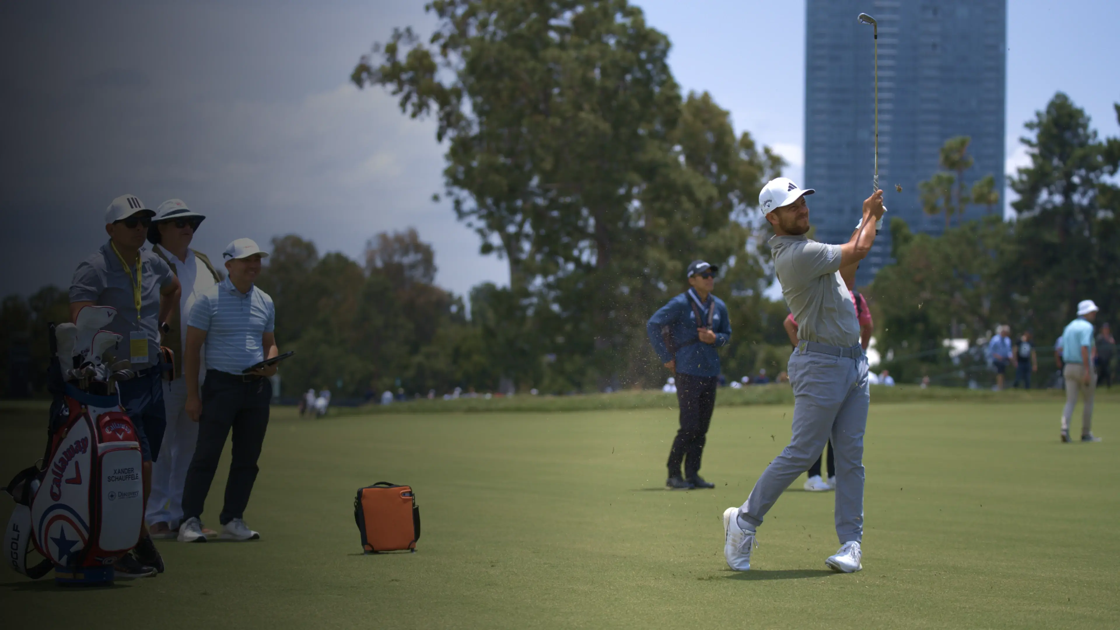 Golfer in mid-swing on a lush green course, with onlookers and trees in the background under a clear sky.